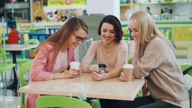 Tri žene sede za stolom u kafiću i gledaju telefon
